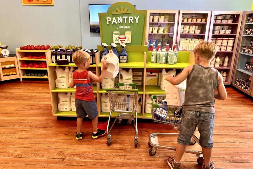 Two boys shop in a pretend grocery store, filling small carts with items from shelves at a museum exhibit. Pensacola Children’s Museum