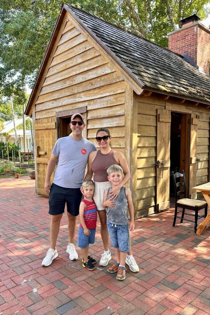 A family of four poses in front of a small wooden house in a historical village, smiling at the camera on a sunny day. Historic Pensacola Village, Florida