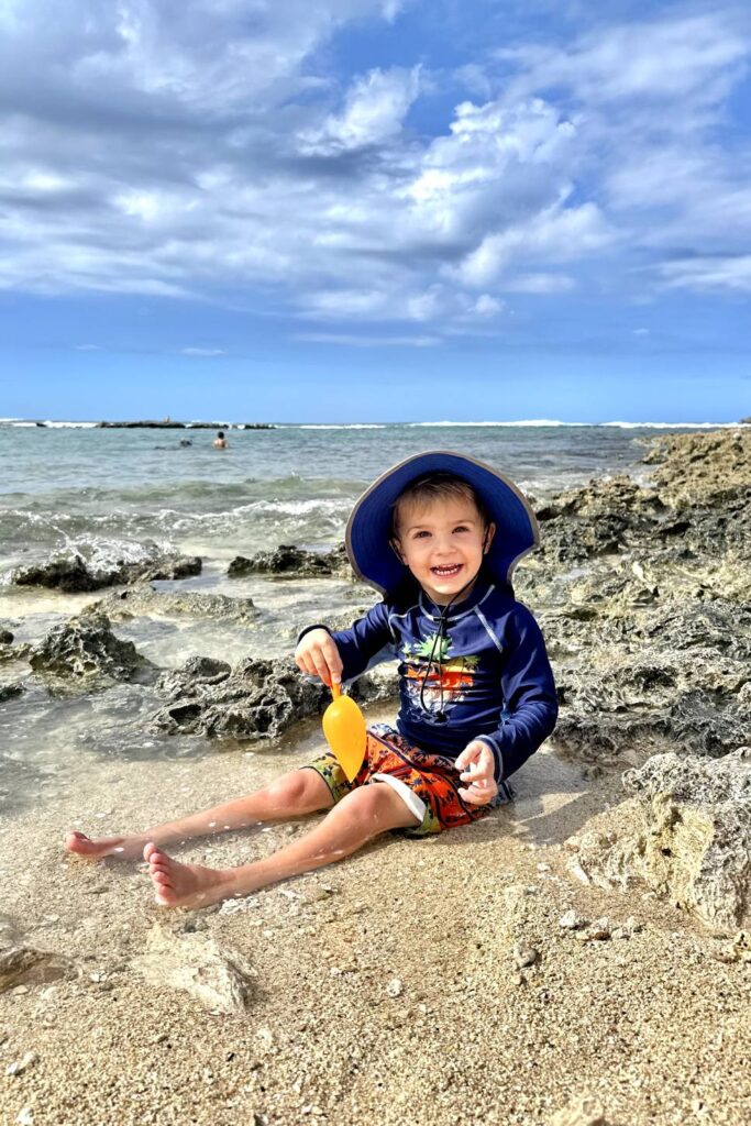 A young boy in swimwear sitting on a rocky shoreline with a shovel, smiling at the camera. Oahu, Hawaii.