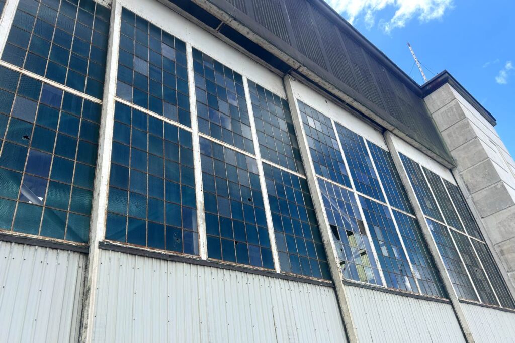 The image shows a large, weathered hangar with tall, blue-tinted windows, some of which are broken or cracked. The building appears aged, with visible signs of wear, and the sky above is clear with a few clouds. The windows' damaged state suggests the hangar's historical significance and age. Aviation Museum in Pearl Harbor, Oahu.