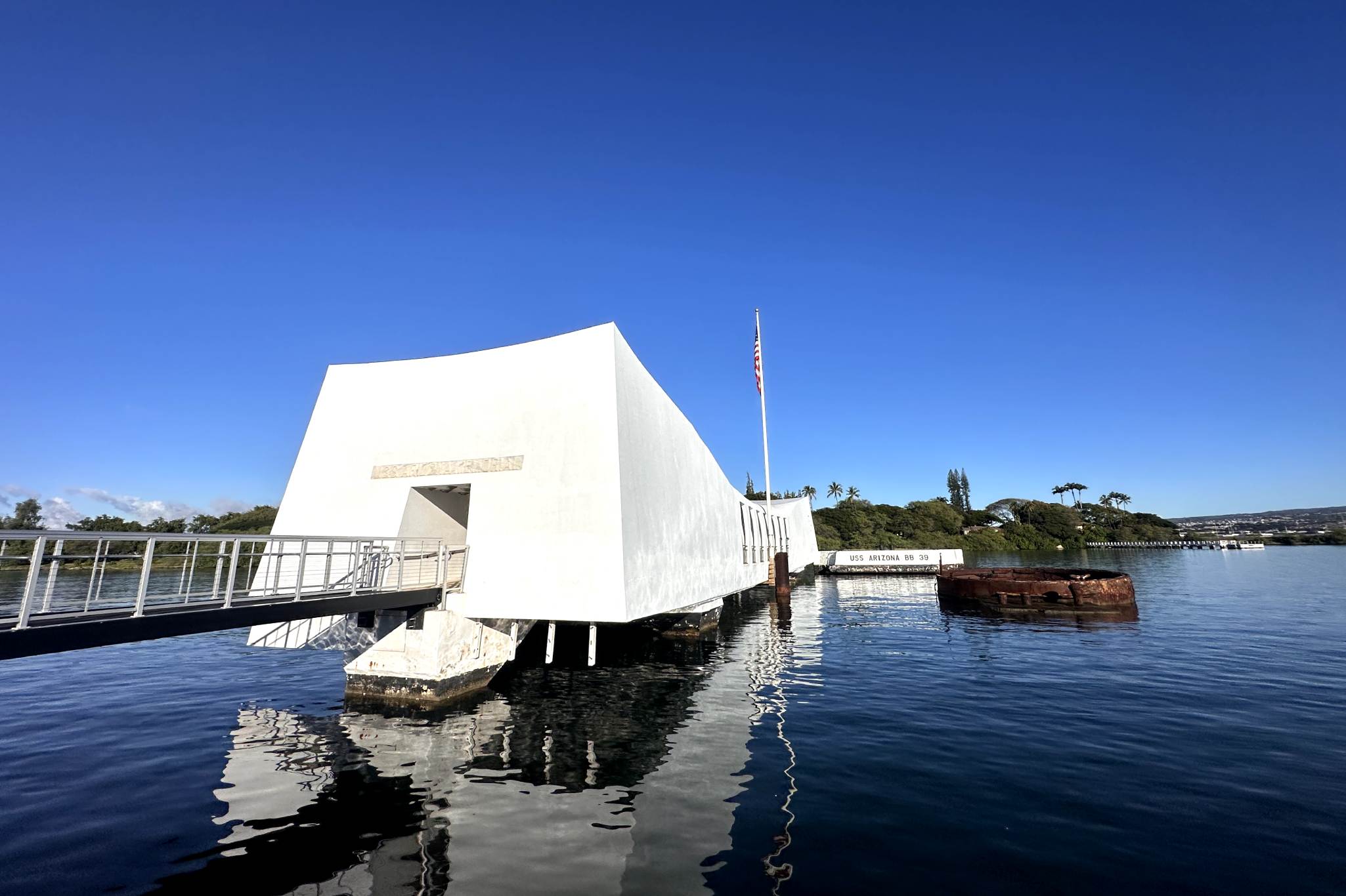 The image shows the USS Arizona Memorial, a white, rectangular structure that spans over the sunken remains of the battleship in Pearl Harbor. An American flag flies at the far end of the memorial, with part of the sunken ship visible in the water nearby. Pearl Harbor, Oahu.