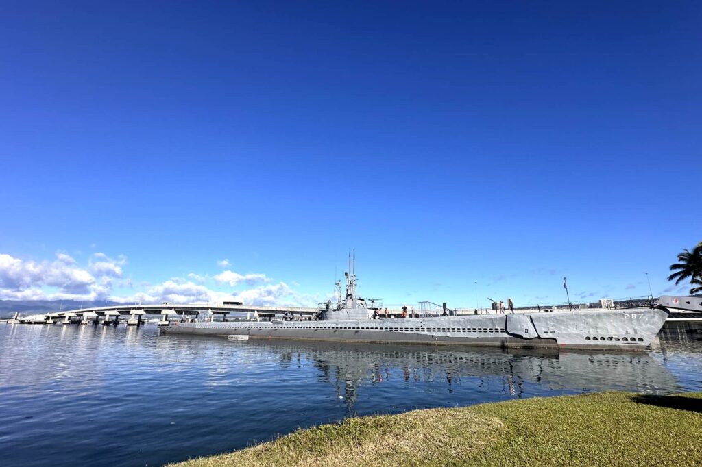 A large, gray submarine is docked in the waters of Pearl Harbor, with a nearby bridge in the background under a clear blue sky. The submarine, identified as "287," is part of a historical exhibit, and a few visitors can be seen walking on its deck. Pearl Harbor, Oahu