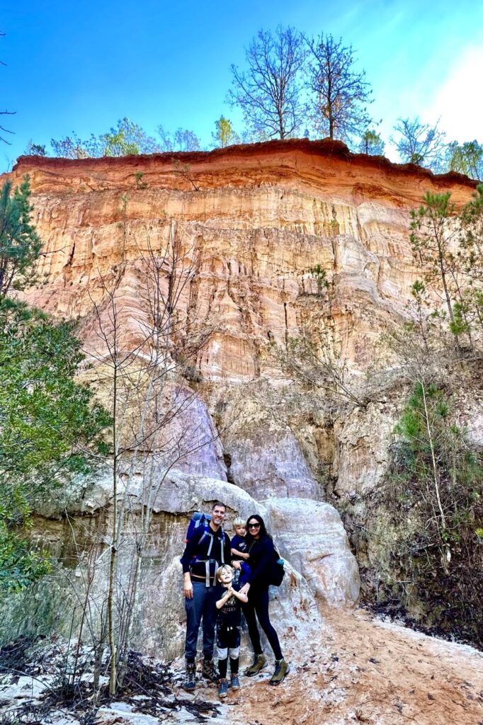 A family of four is standing at the base of a towering, colorful rock formation at Providence Canyon in Georgia. They are smiling, with everyone wearing hiking gear.