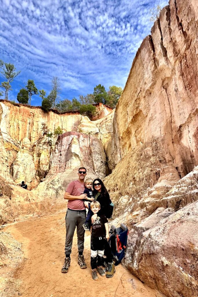 A family of four stands on a sandy trail at Providence Canyon in Georgia, with tall, colorful rock walls rising behind them. They are smiling and dressed for hiking, enjoying their time outdoors under a partly cloudy sky.