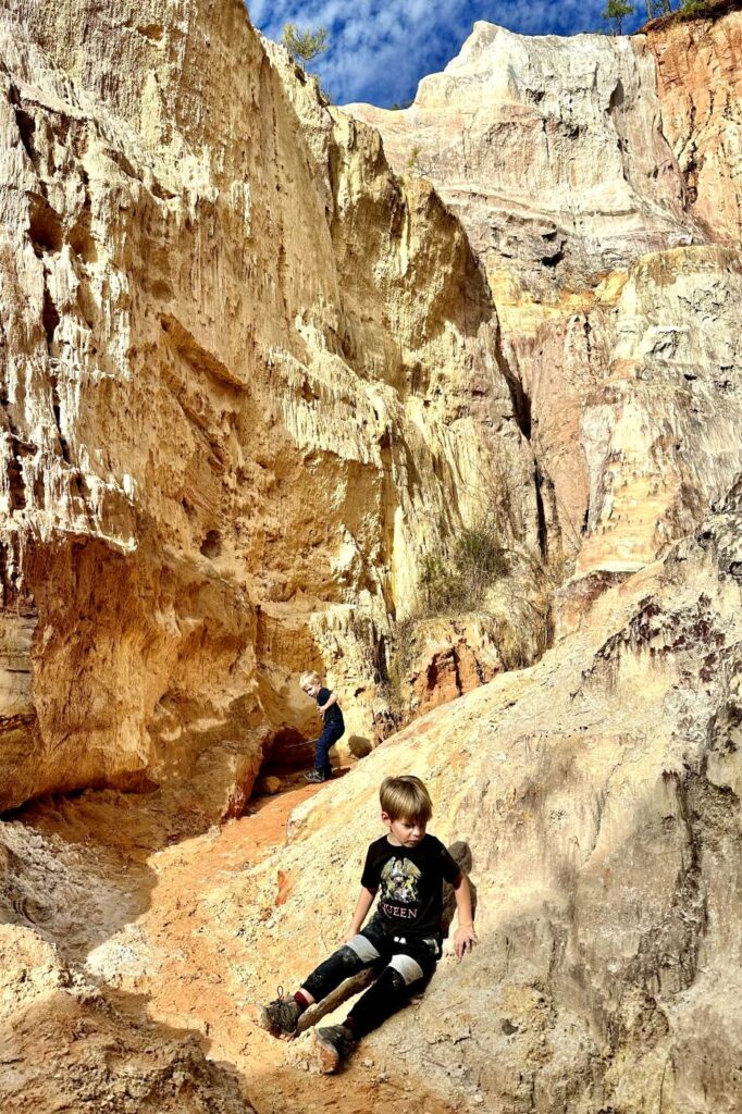 Two children play on the sandy, colorful slopes of Providence Canyon in Georgia, surrounded by tall, layered rock walls in shades of red and tan.