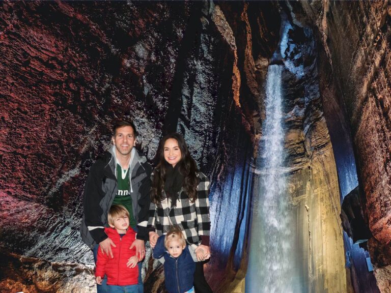 A family of four posing in front of Ruby Falls, with the tall waterfall and colorful lights in the background. Ruby Falls cave walk, Chattanooga, Tennessee