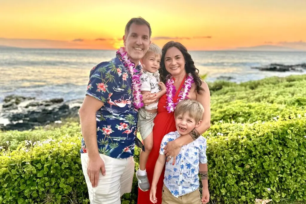 A family of four, dressed in Hawaiian shirts and flower leis, stands together with an ocean sunset behind them, smiling for the camera. Hawaii Family Vacation, Maui, Hawaii.