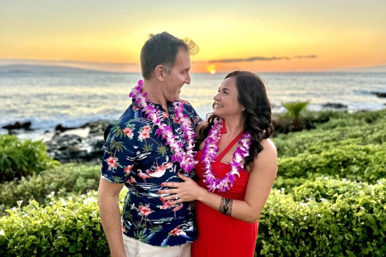 A couple, wearing flower leis, share a warm moment while standing in front of a beautiful ocean sunset. Te Au Moana Luau, Maui, Hawaii.