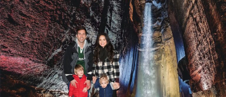 A family of four posing in front of Ruby Falls, with the tall waterfall and colorful lights in the background. Chattanooga, Tennessee