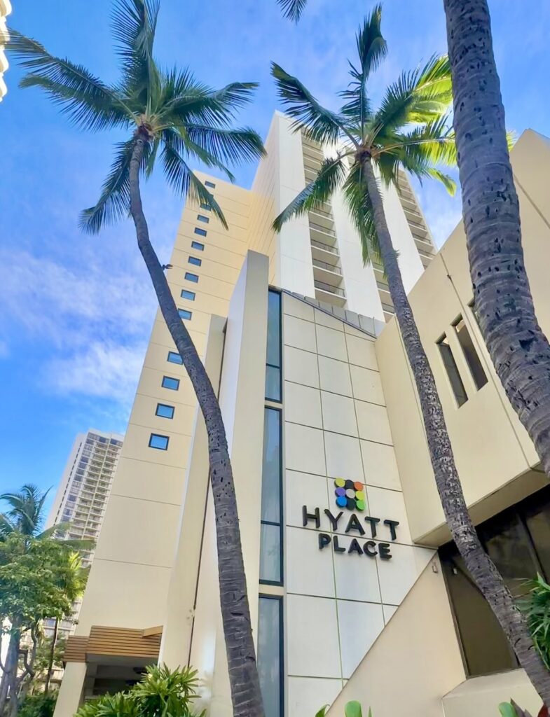 Hyatt Place Waikiki Beach building surrounded by tall palm trees against a bright blue sky.