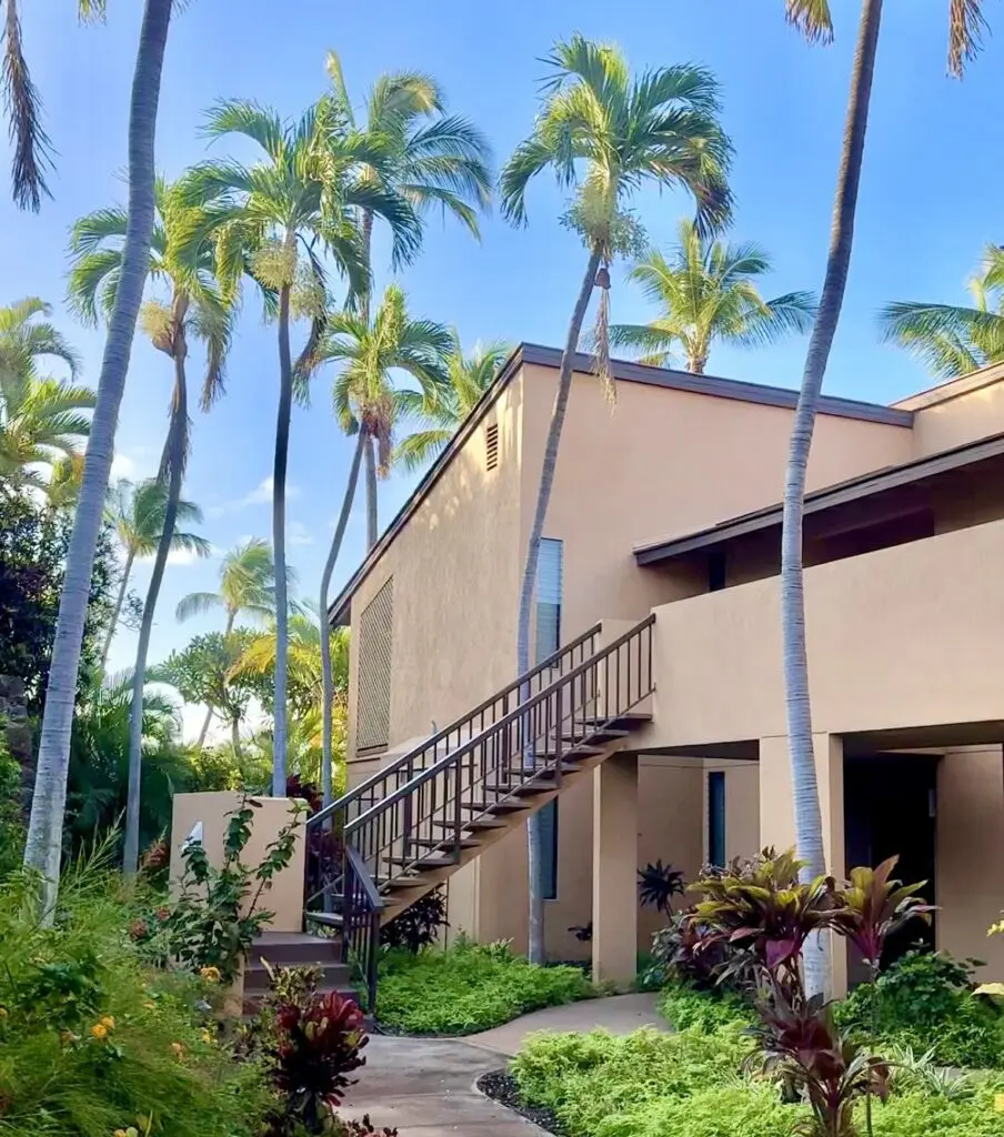 Wailea Ekahi Village condo exterior, surrounded by lush greenery and tall palm trees under a clear blue sky, with a staircase leading to the second floor.