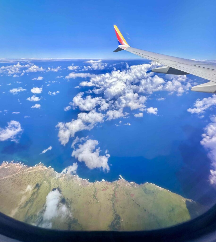 View from a Southwest Airlines plane flying from Oahu to Maui, below a blue ocean, scattered clouds, and the Hawaiian coastline.