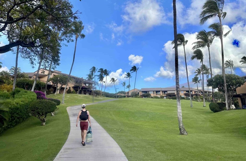 Person walking along a paved path in the grounds of Wailea Ekahi Village under a bright blue sky with scattered clouds.