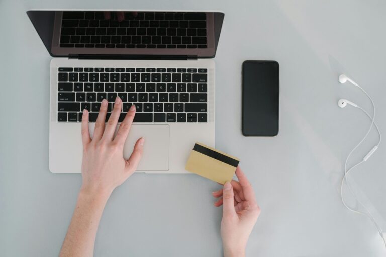 A person on a laptop, typing with one hand and holding a credit card with the other. Nearby, a smartphone, and earphones are placed on a light grey desk. Chase Sapphire Preferred