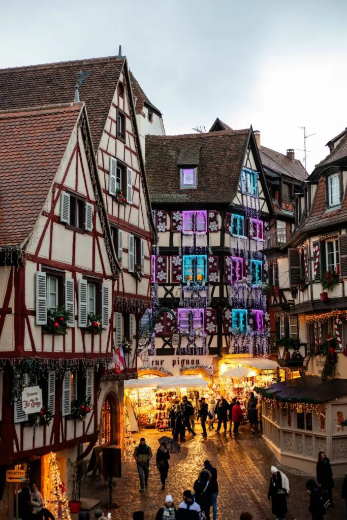 A street view of Colmar, Alsace, with half-timbered houses lit up with Christmas decorations and a festive market scene. Best Christmas Markets in Europe