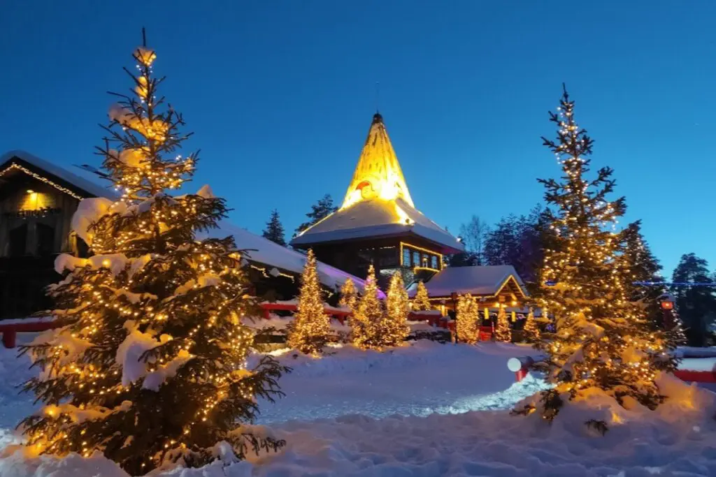 A snowy outdoor scene in Rovaniemi, Finland, featuring a brightly lit Santa Claus Village with Christmas trees and lights.