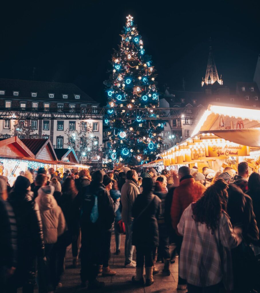 A towering Christmas tree lit with blue and white lights at the center of a Strasbourg Christmas market surrounded by crowds.