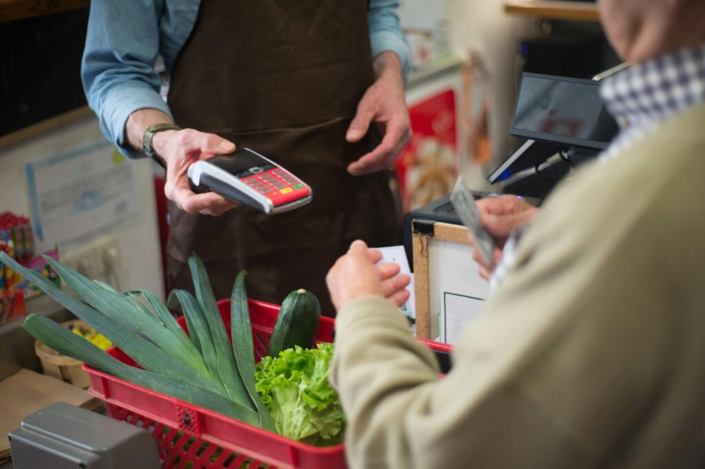 A customer paying for groceries at a store using a contactless card reader, with a basket of fresh vegetables on the counter.