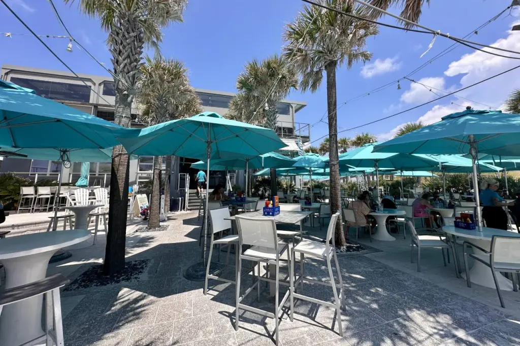 An outdoor dining area in Pensacola featuring bright turquoise umbrellas, palm trees, modern patio furniture, and a relaxed beachside atmosphere under a clear blue sky.