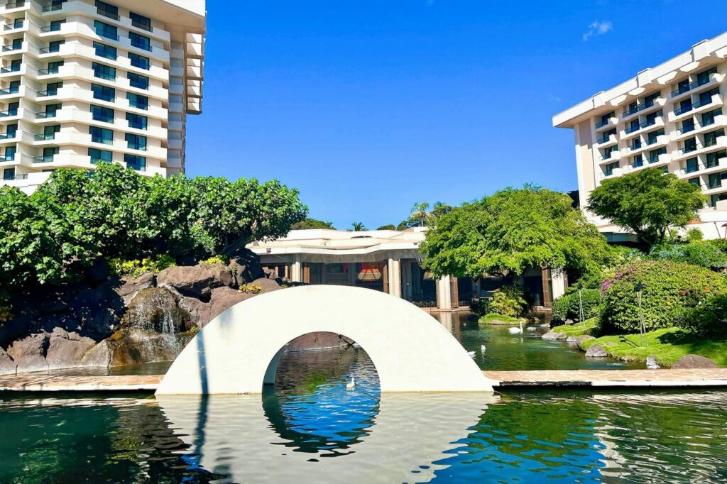 Scenic garden at a Hyatt resort featuring a serene pond with a white arch bridge, and modern hotel buildings under a bright blue sky.