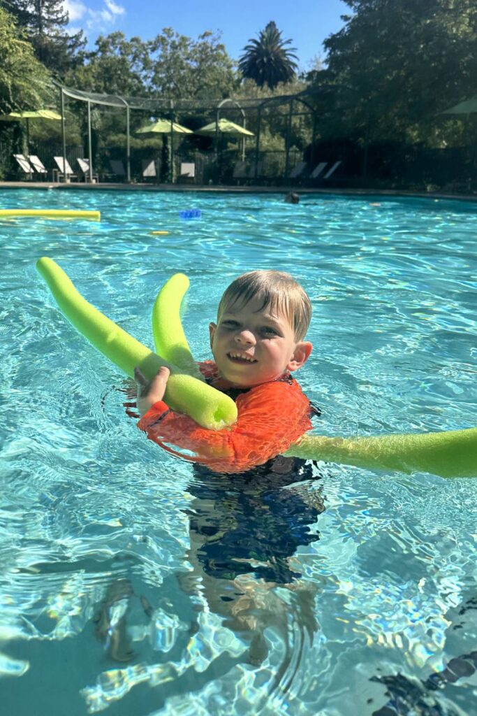 Child enjoying the outdoor pool at a Hyatt hotel, holding pool noodles and smiling under sunny weather.