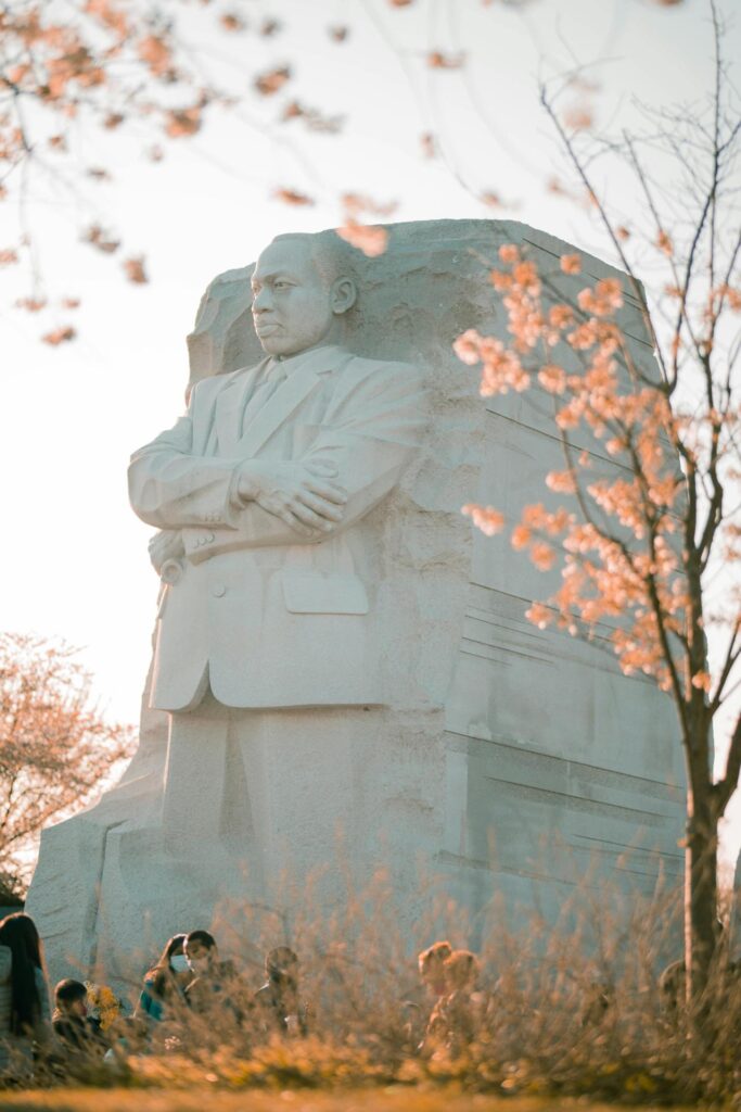 The Martin Luther King Jr. Memorial bathed in warm light, with cherry blossoms framing the statue and visitors walking nearby. Washington DC Cherry Blossoms