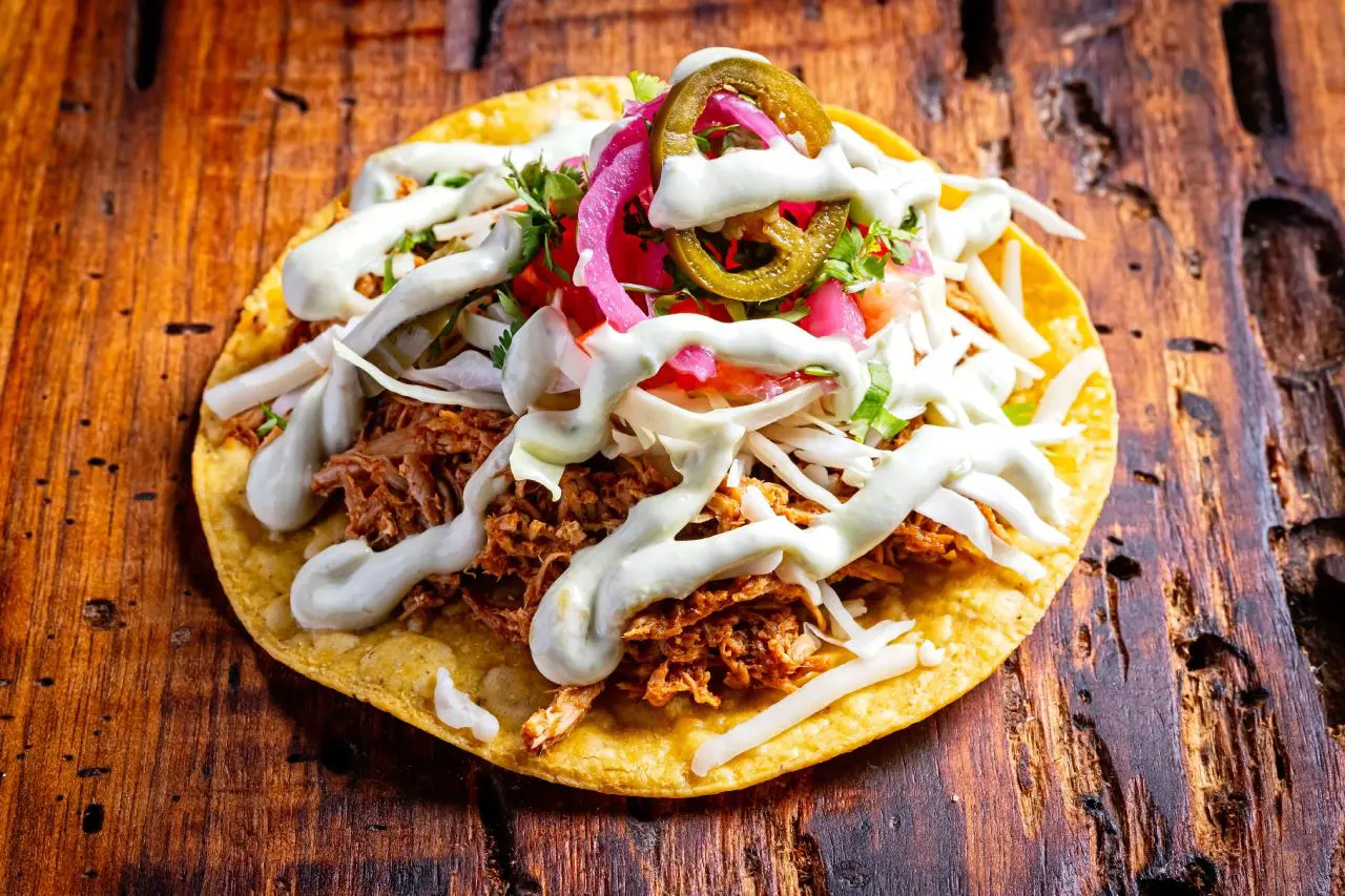 A crispy tostada topped with shredded meat, fresh cabbage, pickled red onions, sliced jalapeños, and drizzled with a creamy white sauce, served on a rustic wooden table.