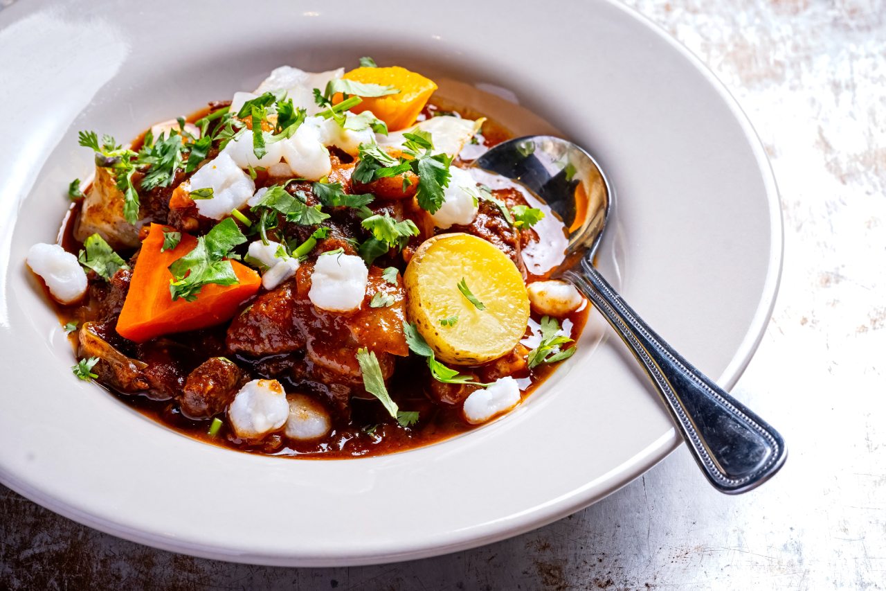 	A white bowl filled with a rich stew containing chunks of meat, potatoes, carrots, white hominy, and garnished with fresh cilantro, with a spoon resting inside.