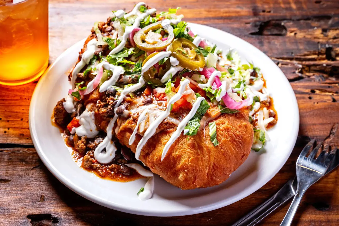 A large, golden-brown fry bread topped with seasoned ground beef, black beans, fresh lettuce, diced tomatoes, pickled onions, jalapeños, and drizzled with a creamy sauce.