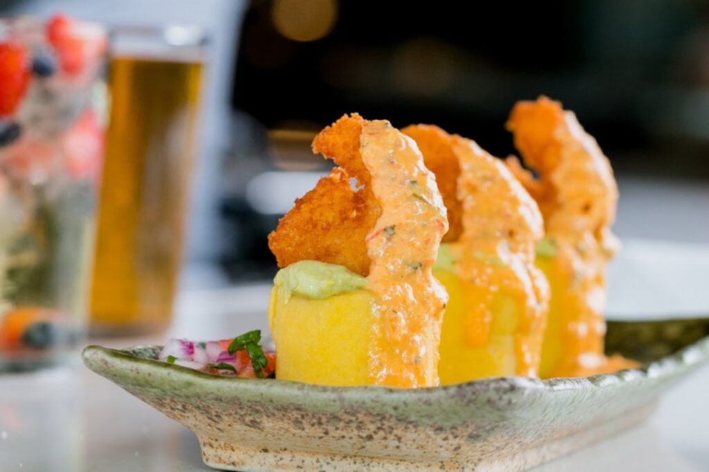 A beautifully plated Peruvian causa dish featuring layers of yellow mashed potatoes topped with creamy avocado, crispy golden shrimp, and drizzled with a rich, spicy orange sauce, served on a rustic ceramic plate with a side of fresh salsa.