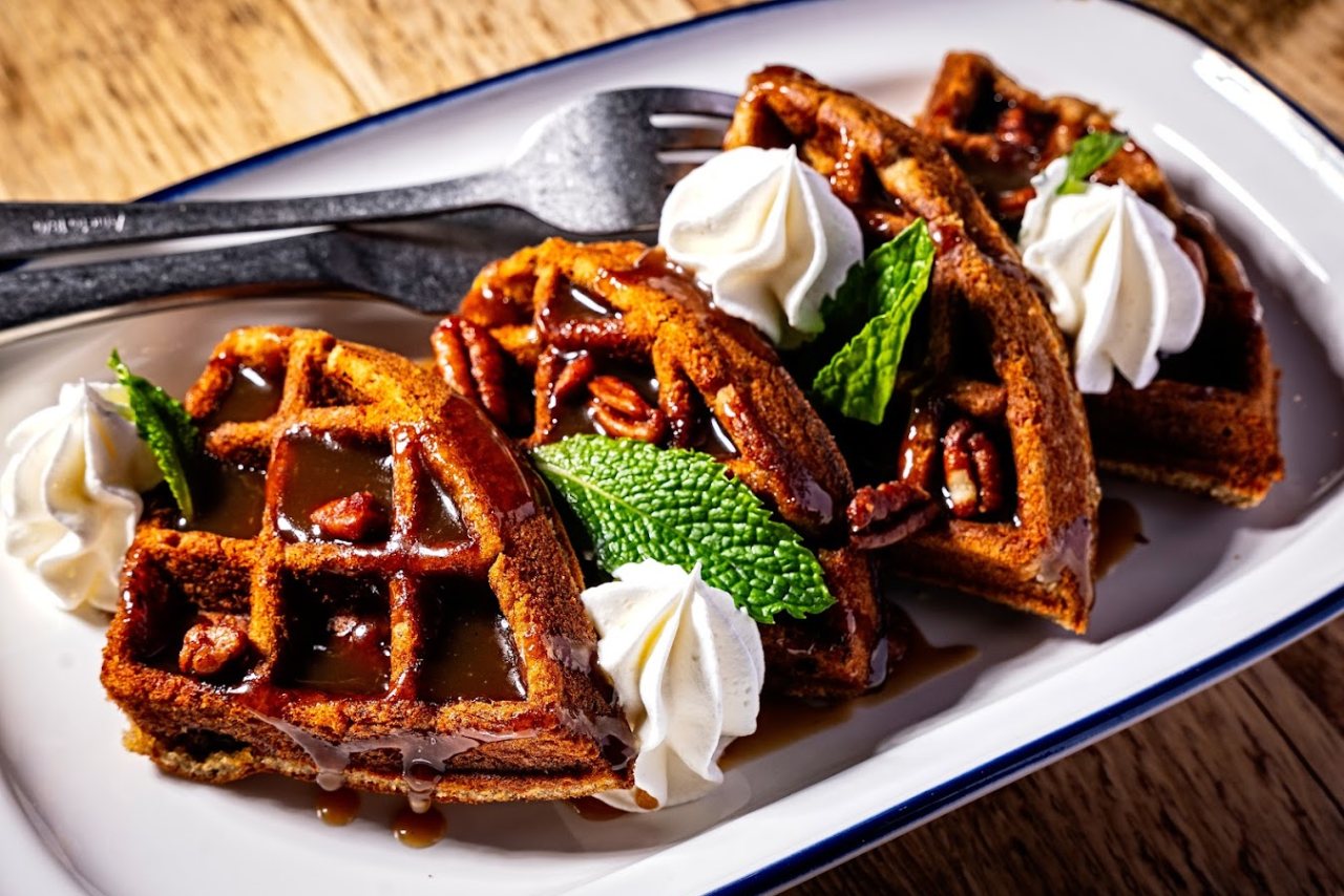 A plate of golden-brown waffles topped with caramelized pecans, drizzled with rich syrup, and garnished with fresh mint leaves and whipped cream, served on a white dish with a fork. 