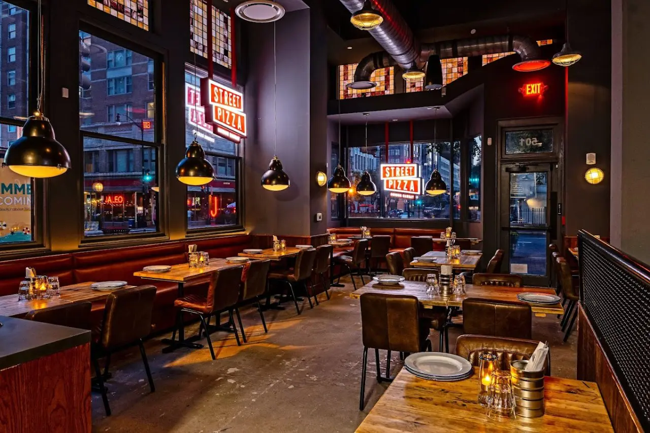 Gordon Ramsay Street Pizza restaurant interior with dim lighting, wooden tables, and leather chairs. Large windows display a city street with a neon sign reading "Street Pizza."