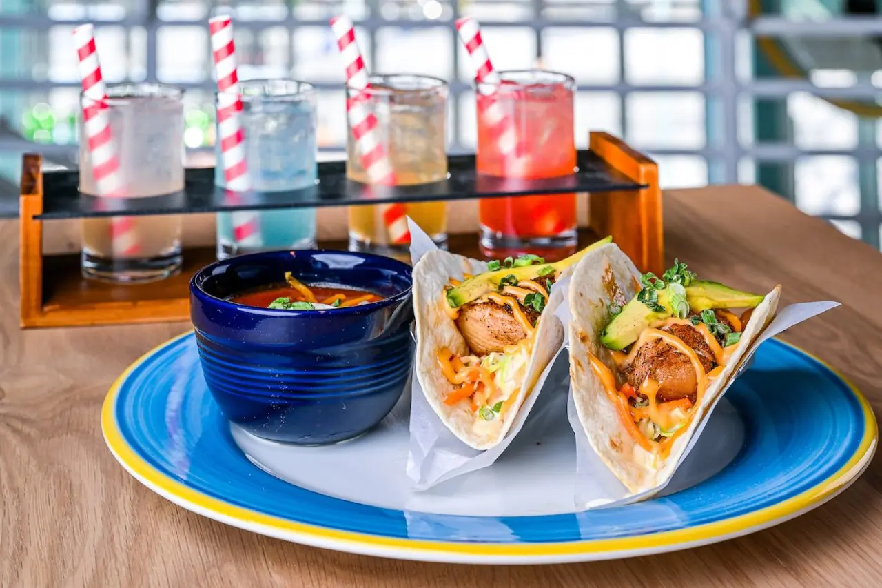 A colorful plate featuring two grilled fish tacos topped with avocado slices, shredded vegetables, and drizzled with a creamy orange sauce, served alongside a bowl of soup. Boardwalk Bar & Arcade