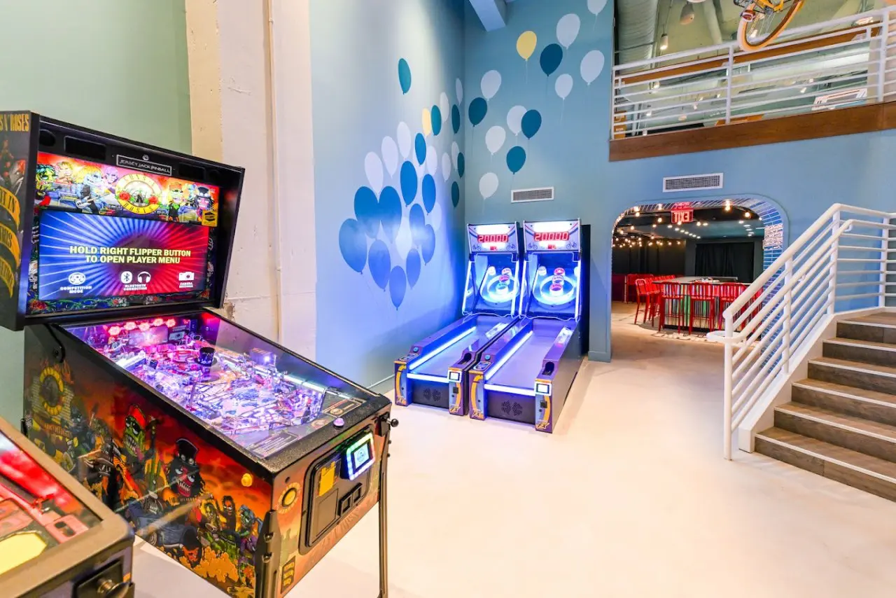 A lively arcade area with a black and red pinball machine and two glowing blue skee-ball games. The walls are decorated with balloon graphics, and a staircase leads to an upper level. 