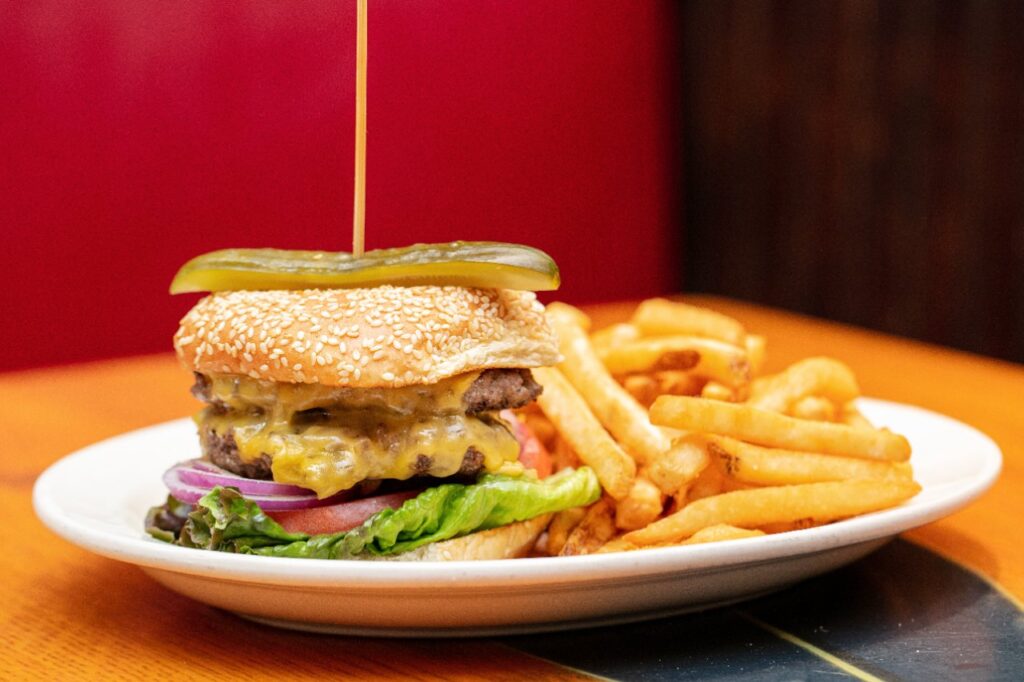 A juicy double cheeseburger with melted cheese, fresh lettuce, tomato, red onion, and a sesame seed bun, topped with a pickle and held together by a skewer. The Tombs