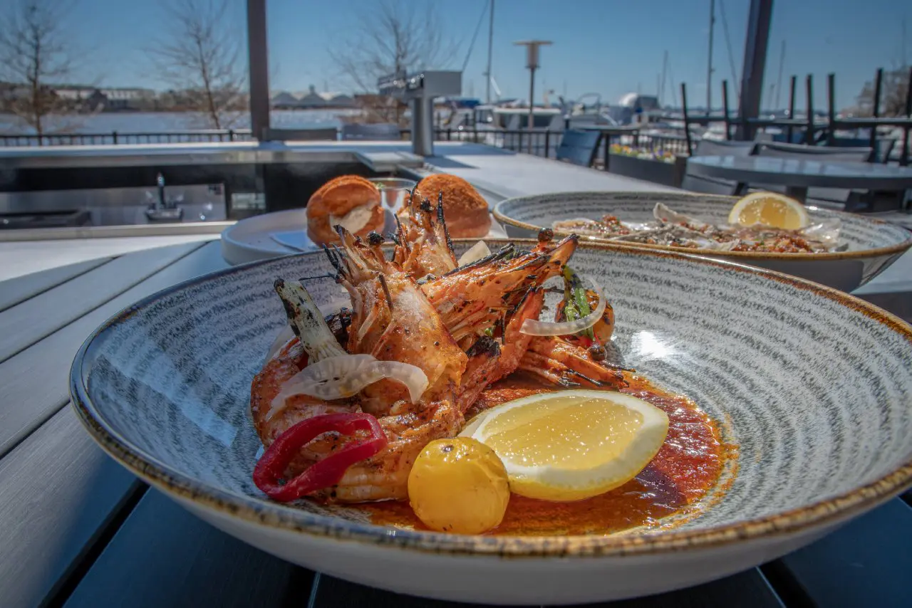 A bowl of grilled prawns with slightly charred shells, served in a rich, red sauce with sliced onions, peppers, and a lemon wedge. 