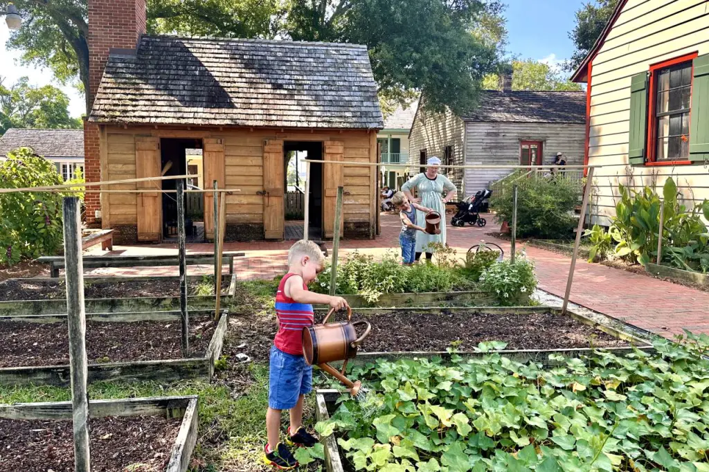 A young boy watering plants in a raised garden bed, supervised by a woman in period clothing in a historical village. Pensacola, Florida.