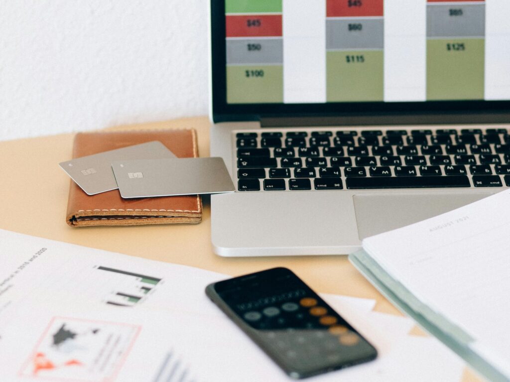 Two business credit cards sit on a wallet next to a laptop with graphs on the screen. Below them are more paper graphs and an iPhone showing the calculator app.