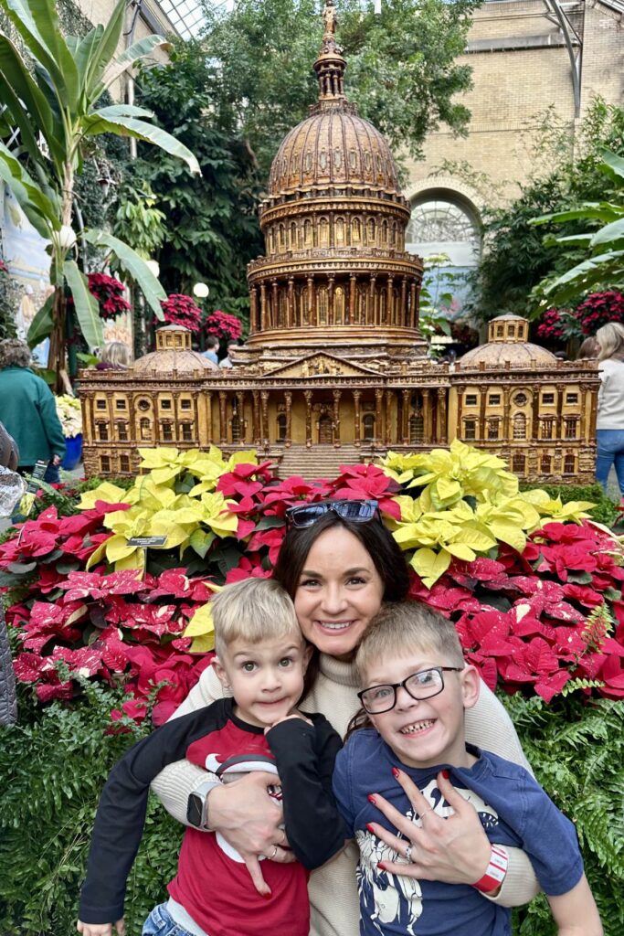 A mother and her two young sons smiling in front of a model of the U.S. Capitol surrounded by poinsettias. US Botanical Gardens.