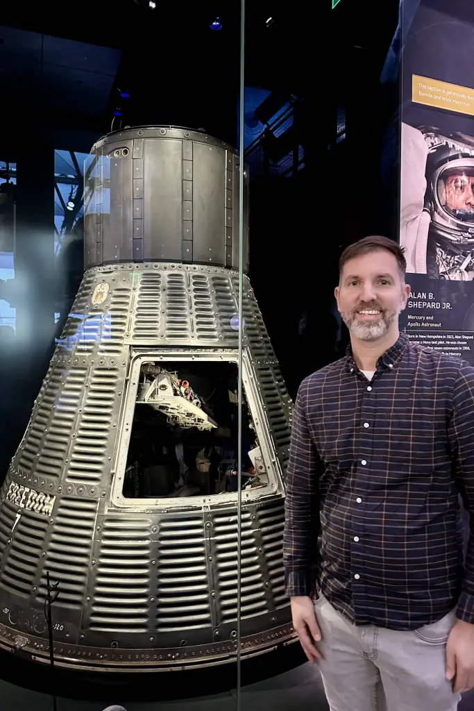 A man standing next to a Mercury spacecraft exhibit inside a museum, with a plaque showing details about astronaut Alan B. Shepard Jr. in the background. Air and Space Museum. Washington DC Itinerary