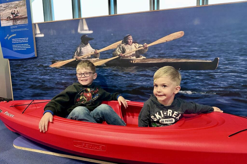 Two boys seated in a red kayak in an interactive exhibit simulating traditional boat paddling on the water.  National Museum of the American Indian