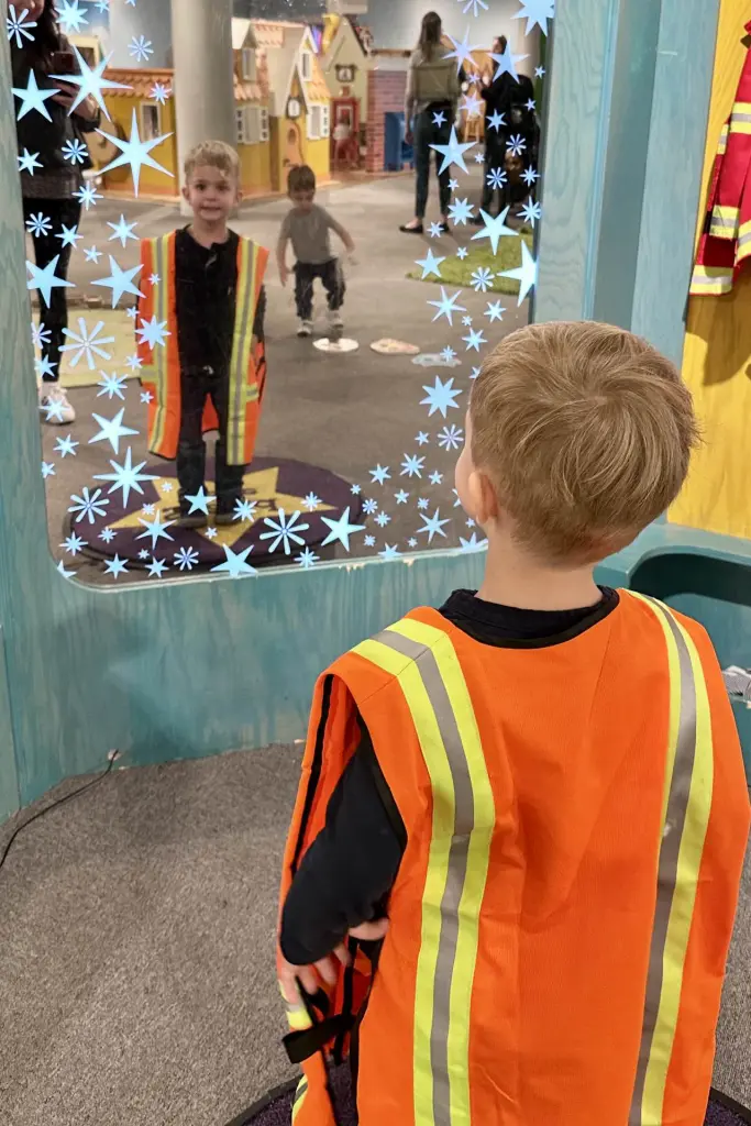 A young boy dressed in a bright orange safety vest, looking into a mirror with star patterns. National Children’s Museum