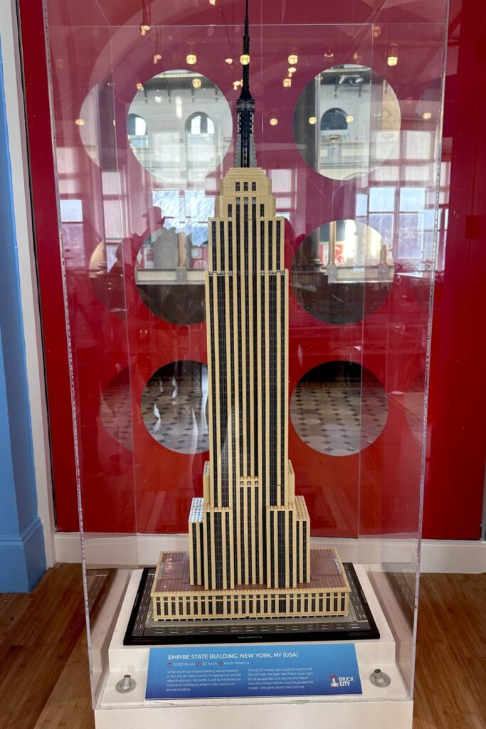 LEGO model of the Empire State Building in a glass case, with a plaque stating it has 15,200 bricks and took 65 hours to build. The background features a red wall with circular cutouts and exhibit reflections.