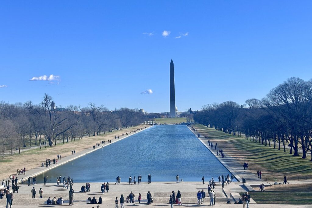 The Washington Monument viewed across the Reflecting Pool, surrounded by visitors and trees.