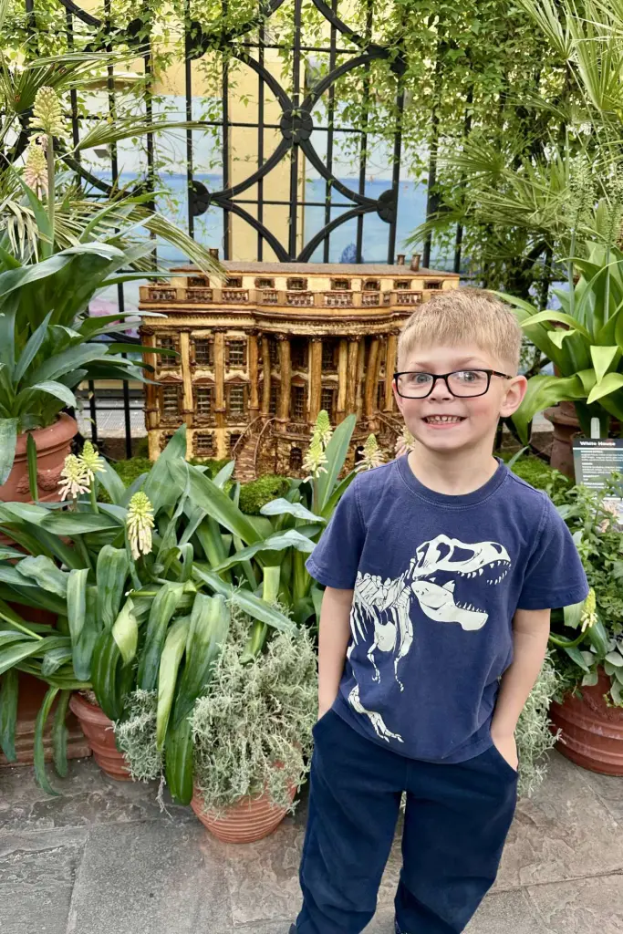 A young boy, wearing a blue dinosaur t-shirt, stands smiling in front of a detailed miniature model of the White House made from natural materials, surrounded by green plants. US Botanical Gardens