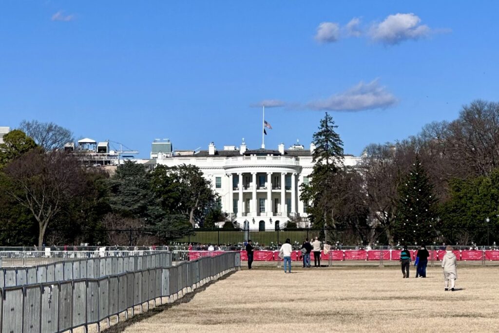 A view of the White House with a bright blue sky, a half-mast American flag flying on top, and a large Christmas tree on the right. A few visitors stand behind a fenced-off area, taking photos.