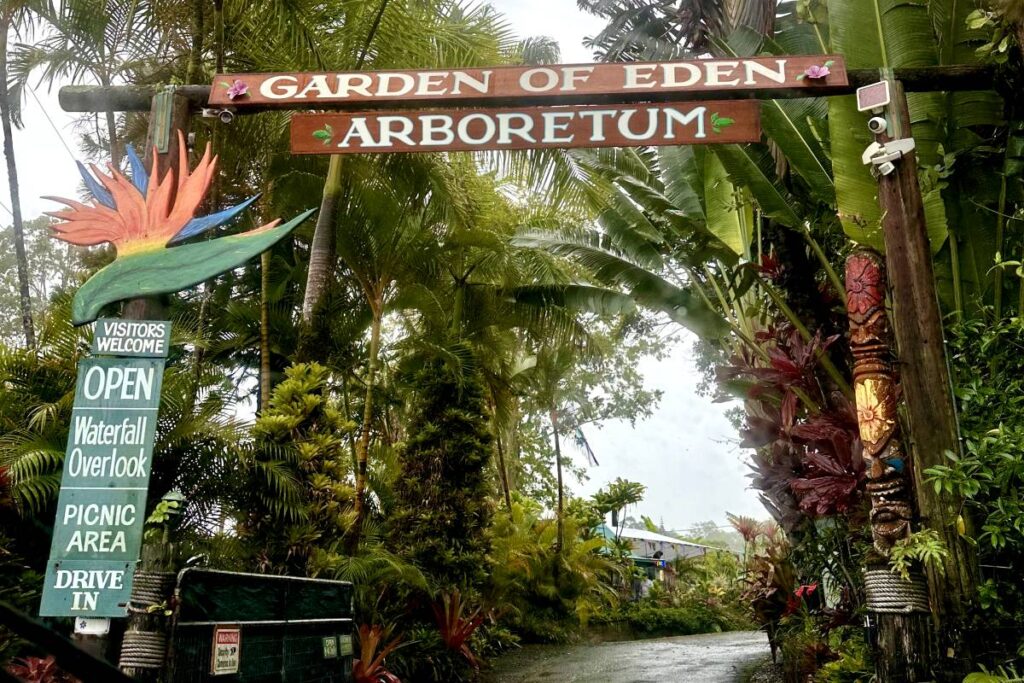 Entrance to the Garden of Eden Arboretum with a large wooden sign above the pathway, surrounded by tropical plants and a bird of paradise sculpture.