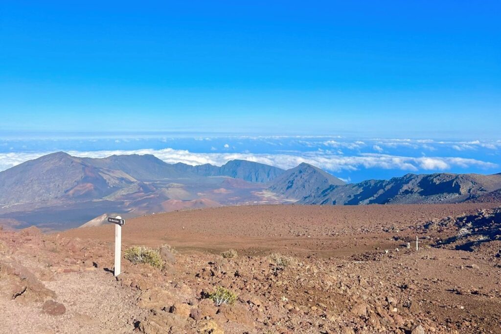 A panoramic view of Haleakalā National Park’s crater, featuring barren, rocky terrain with faint walking trails and mountain ranges in the distance.