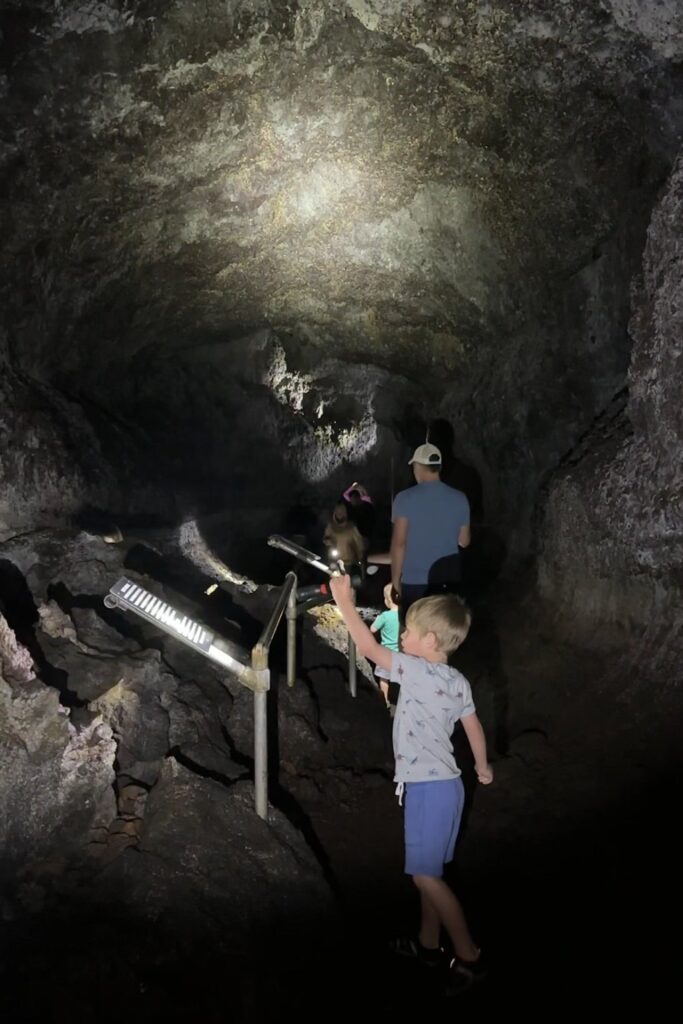 A family walks deeper into the dimly lit lava tube cave, with flashlight beams illuminating the rocky walls and uneven path.