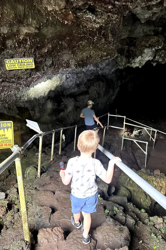A child descends a rocky path inside the lava tube, gripping the metal handrails while navigating the cave's uneven terrain.